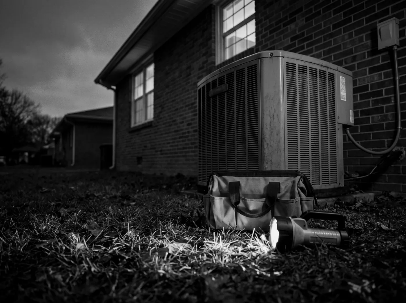 A tool bag and flashlight beside an outdoor air conditioning unit at dusk