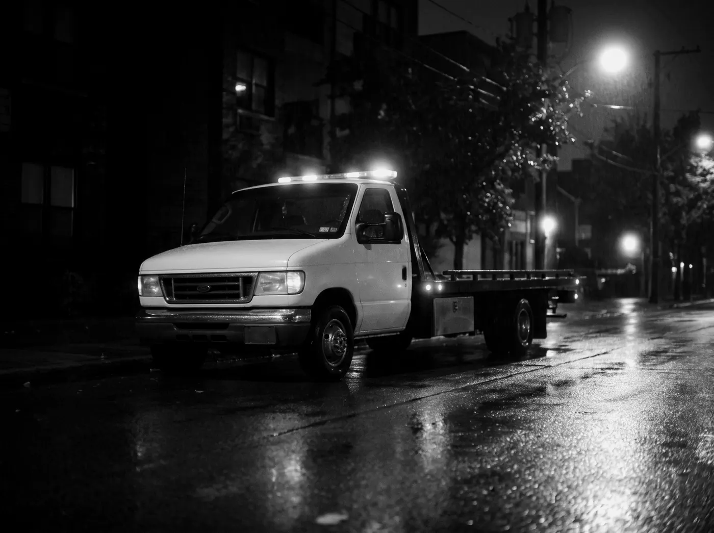 A white tow truck with amber lights on a wet street at night